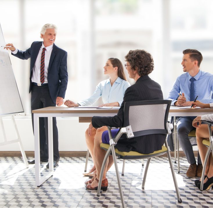 Positive senior leader standing at flipchart at meeting at drawing graph to his team. Multiethnic business people looking at him and smiling